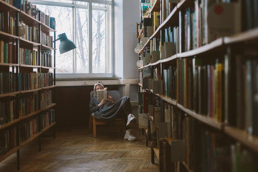 A woman engrossed in reading a book amidst the serene ambiance of a library. Soft natural light illuminates her surroundings, as she focuses on the pages in her hands, lost in the world of literature."